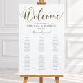A wedding welcome sign printed on a white board with elegant gold script and black table seating details, displayed on a wooden easel in a bright indoor setting.
