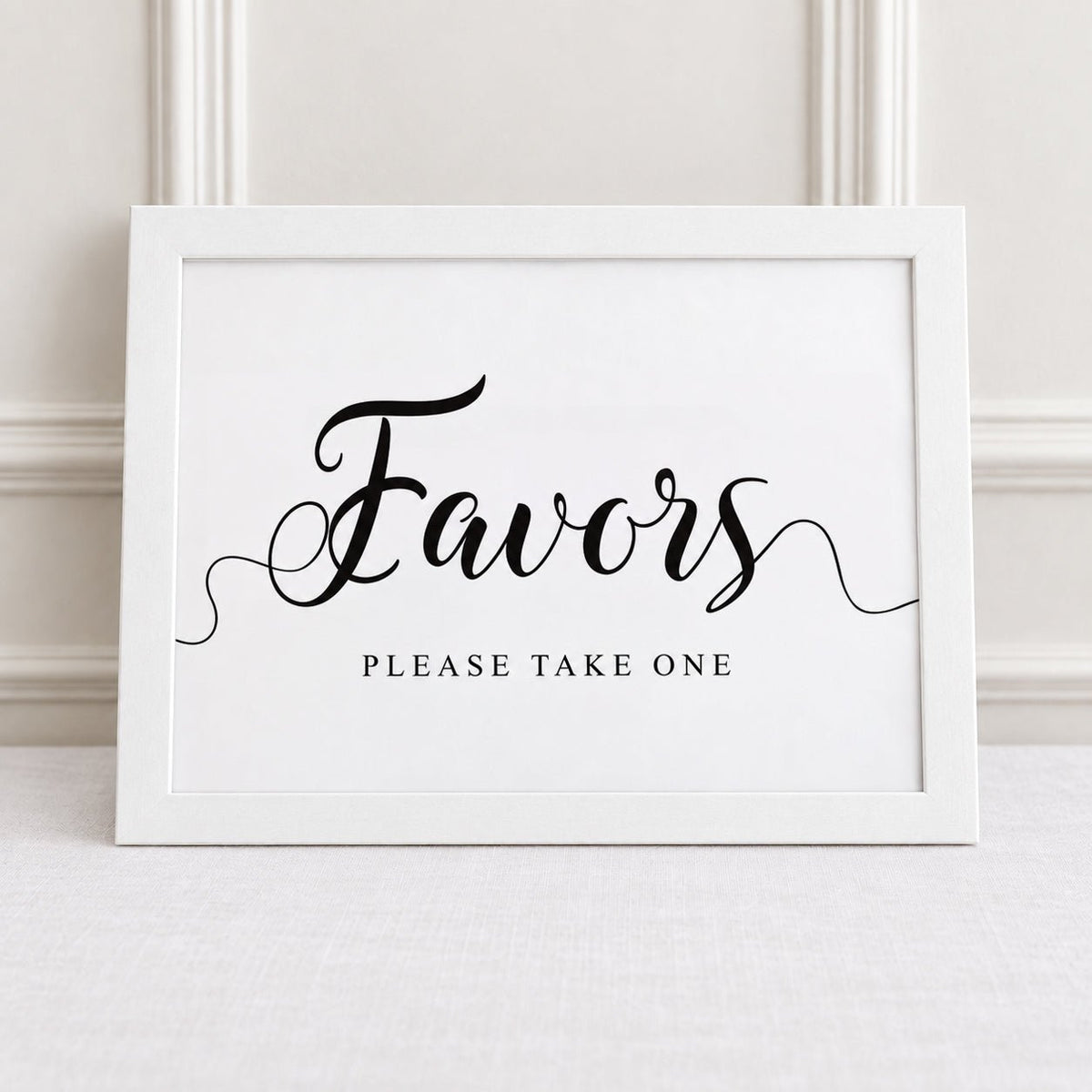 A white framed sign with black script reading 'Favors' and smaller text 'PLEASE TAKE ONE' below, displayed against a light neutral wall and surface.