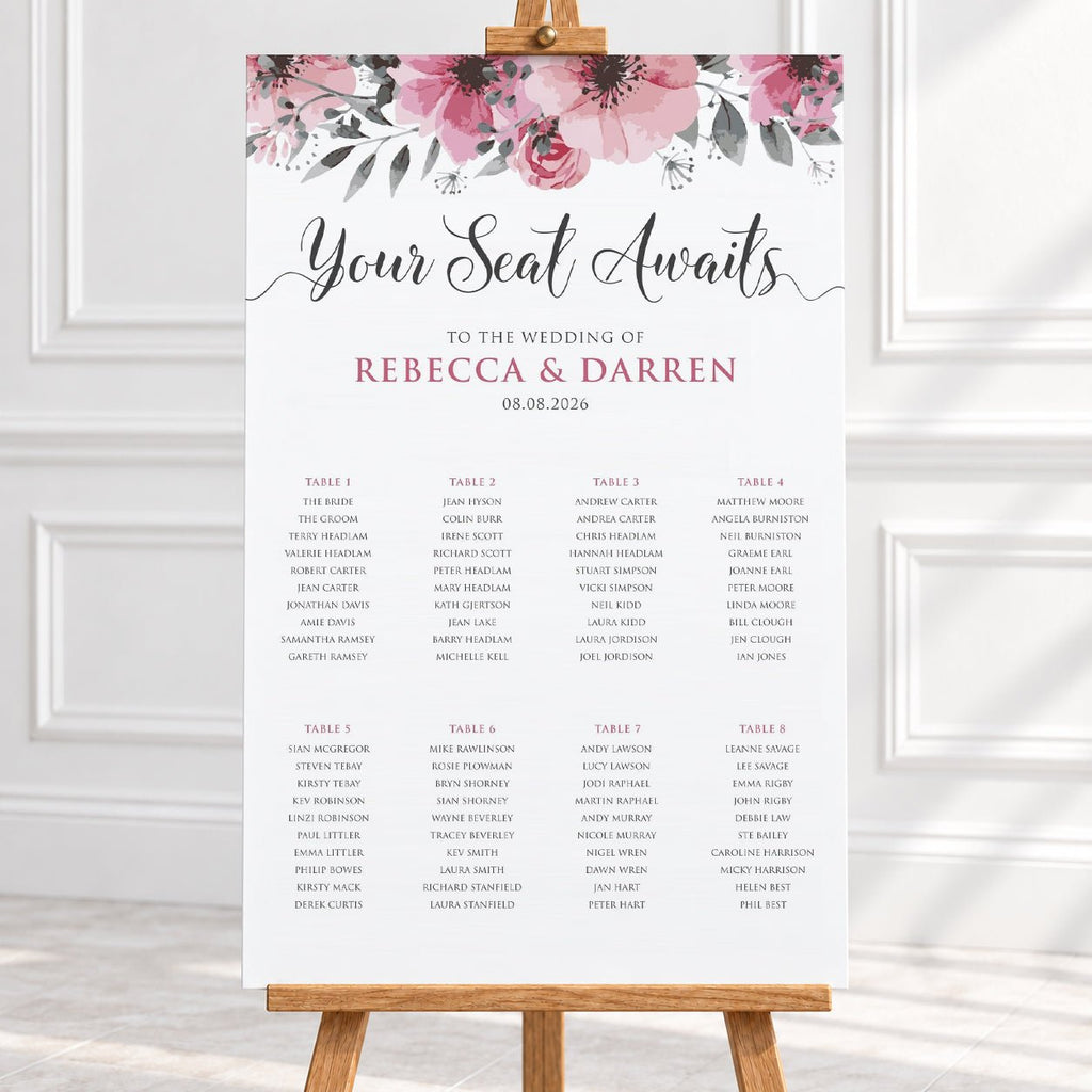 A wedding seating chart print with a pink floral watercolor header and elegant script reading 'Your Seat Awaits' above tables of names, displayed on a wooden stand against a white wall and light fabric background.
