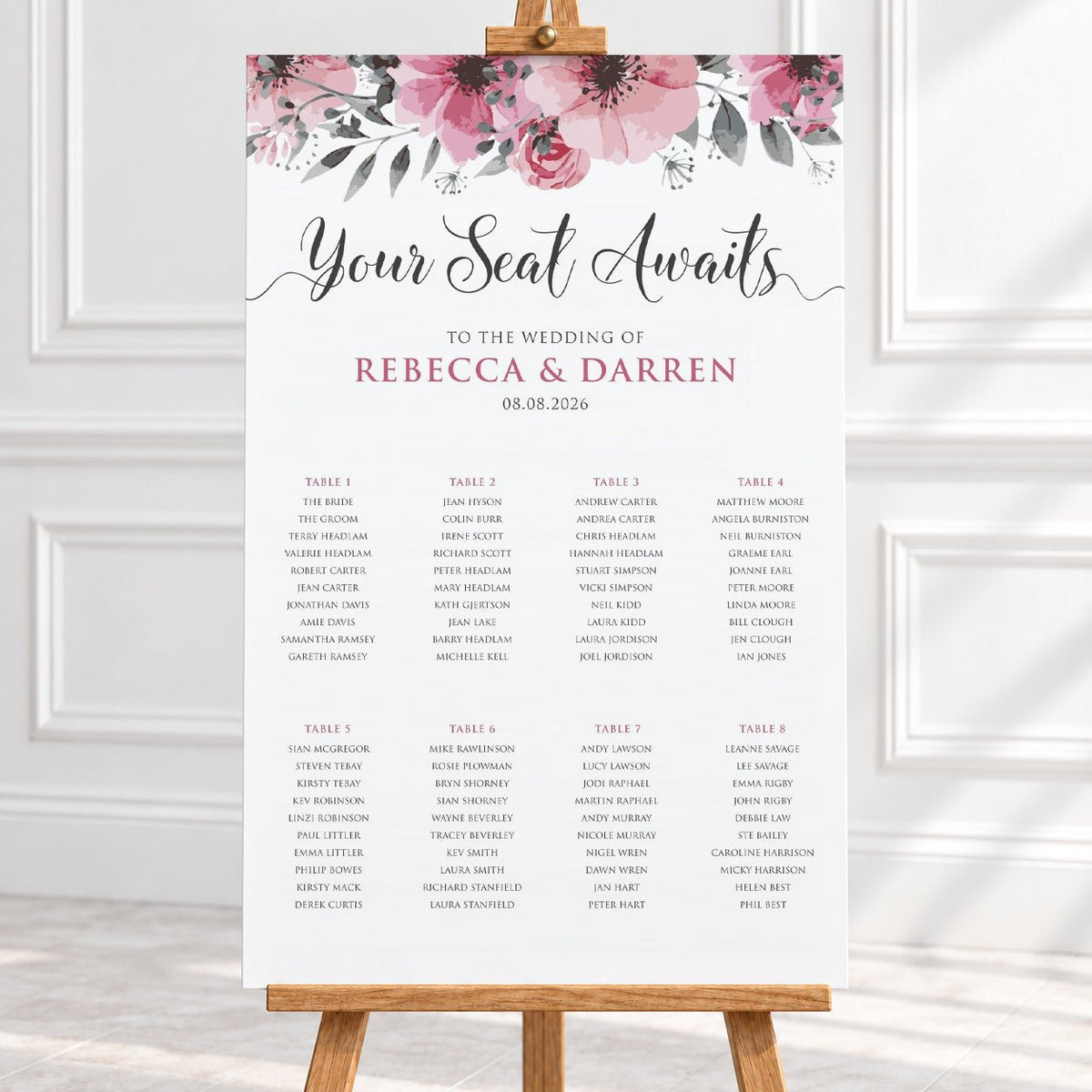 A wedding seating chart print with a pink floral watercolor header and elegant script reading 'Your Seat Awaits' above tables of names, displayed on a wooden stand against a white wall and light fabric background.