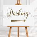 A white rectangular sign with the word 'Parking' in large brown script and a matching right-pointing arrow, displayed on a wooden easel in a bright indoor setting.