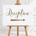 A white reception sign with elegant gold script reading 'Reception' and a matching gold arrow pointing right, displayed on a wooden easel in a bright indoor setting.