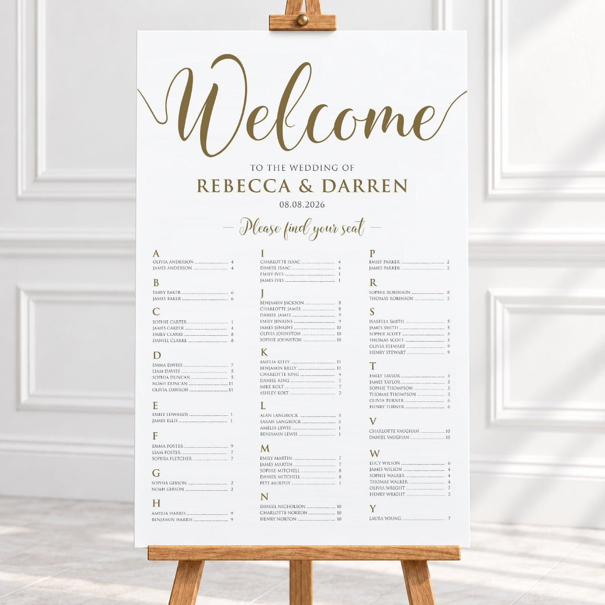 A wedding welcome sign displayed on a wooden easel in a bright interior. The sign is white with elegant gold script reading 'Welcome' and gold personalized text for a wedding of Rebecca & Darren, with a seated guest list printed below.