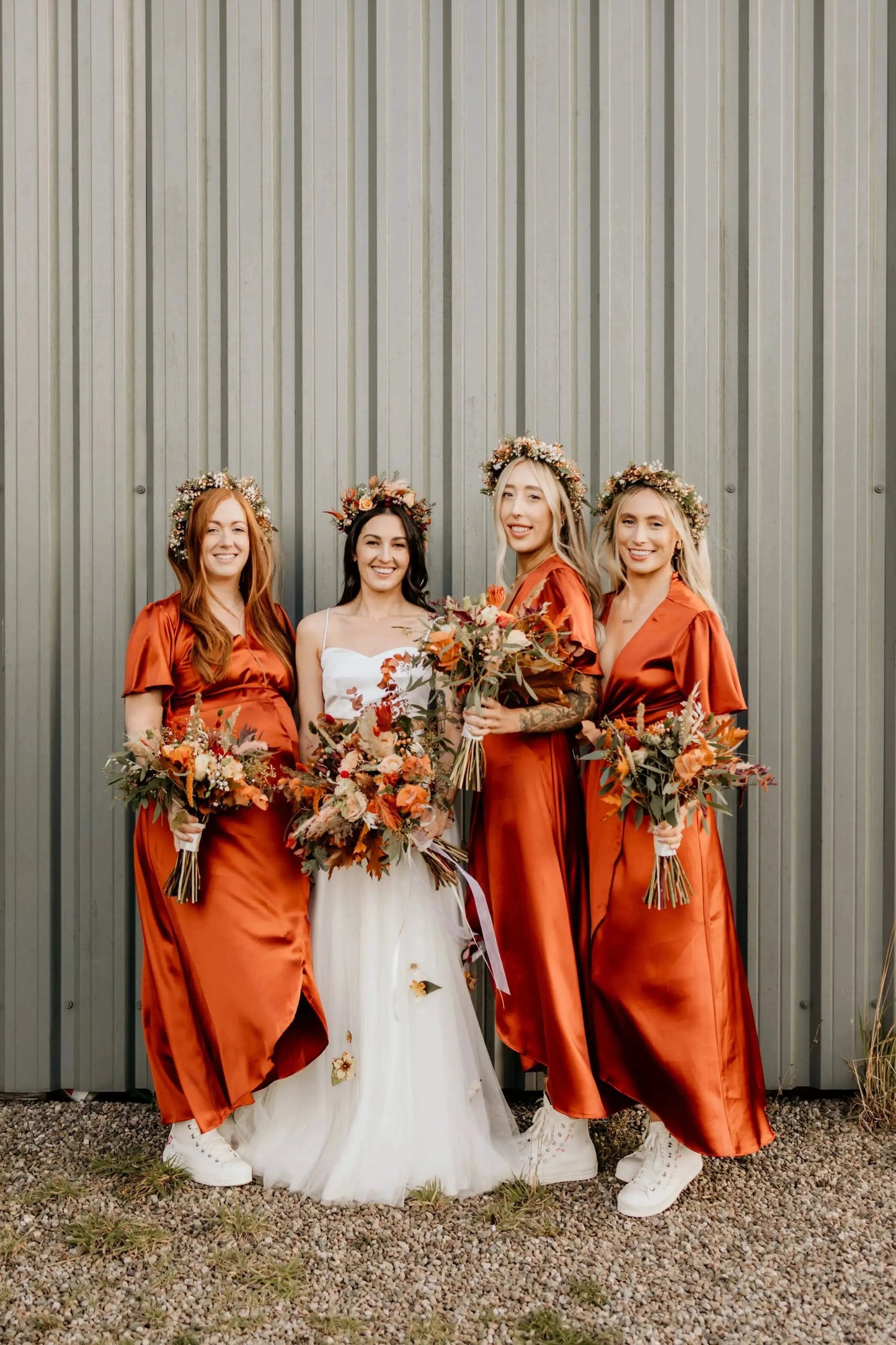 Bride and three bridesmaids in burnt orange bridesmaid dresses standing in front of a metallic wall.