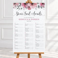 A wedding reception seating chart printed on a large white board with pink floral decoration at the top, displayed on a wooden easel in a bright indoor setting.