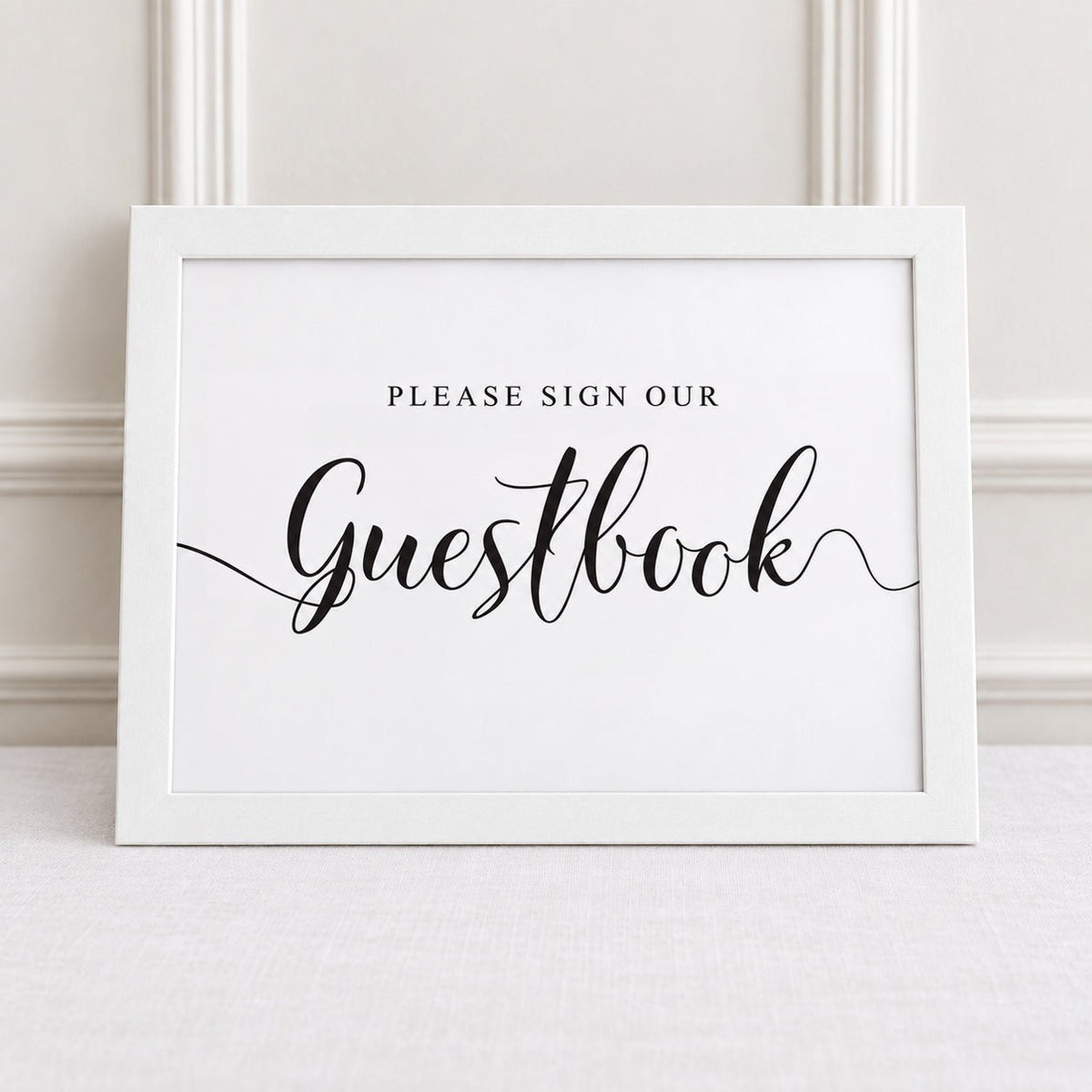 A white framed guestbook sign with black text reading 'PLEASE SIGN OUR Guestbook' displayed against a light beige paneled wall and textured fabric surface.
