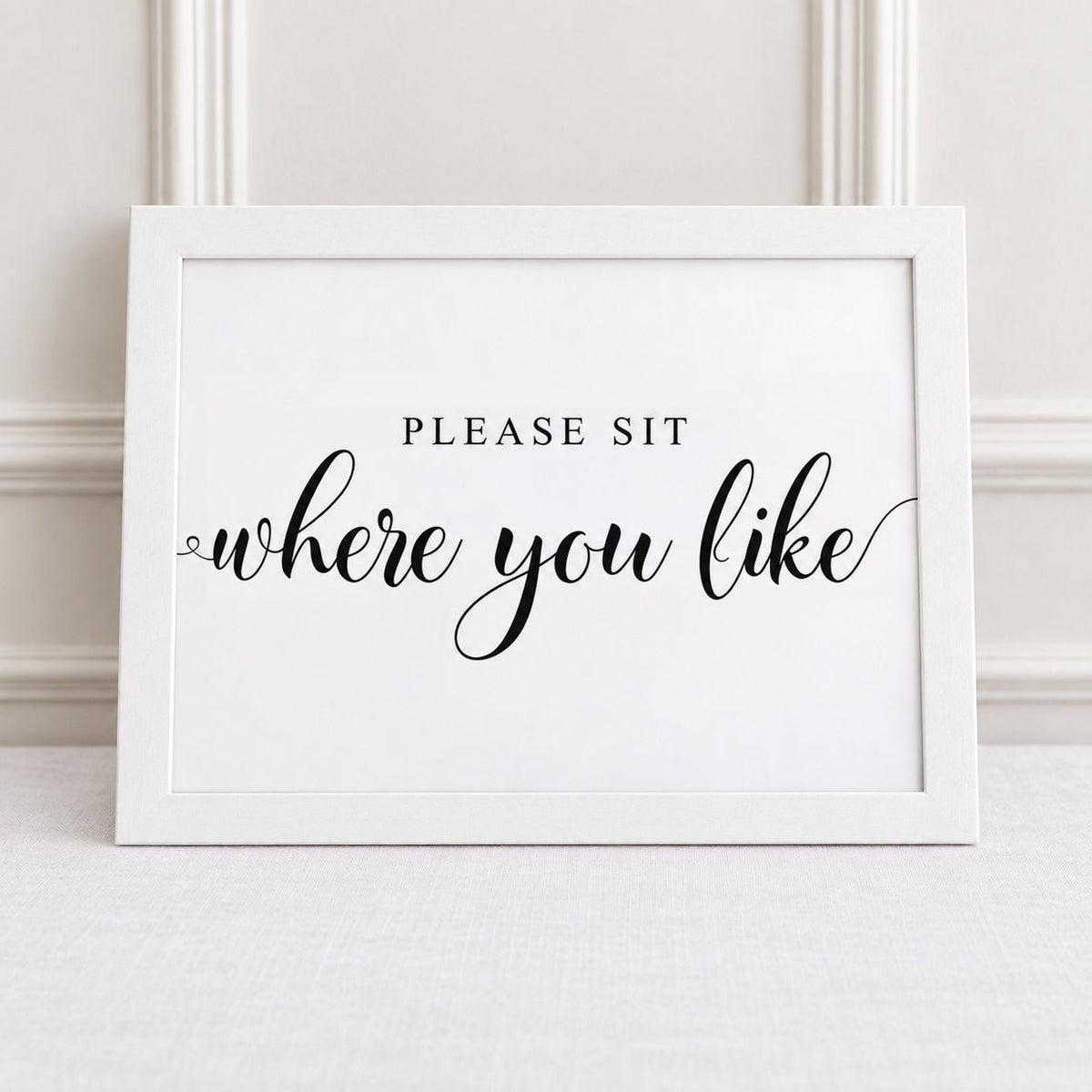 A white framed typographic wall sign reading 'PLEASE SIT where you like' in black lettering, leaning against a light neutral wall on a pale surface.