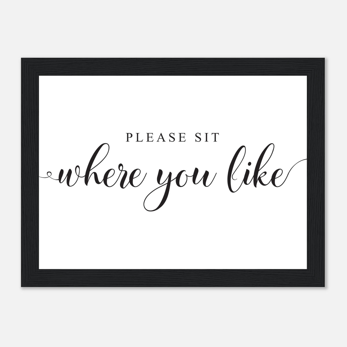 A framed typographic wall sign with the phrase 'PLEASE SIT where you like' in black lettering on a white background, enclosed in a black frame against a light gray backdrop.