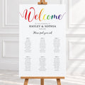 A wedding welcome sign displayed on a wooden easel. The sign features a colorful gradient 'Welcome' script at the top and a seated guest list printed below in black text on a white background.