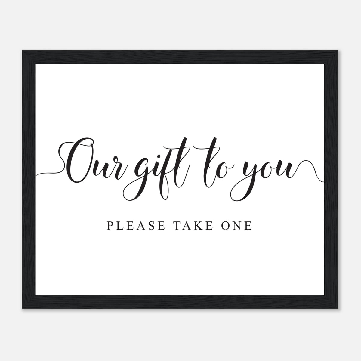 A framed typographic wall sign with a black frame, white background, and black text reading 'Our gift to you' in script with 'PLEASE TAKE ONE' below, shown on a light gray background.