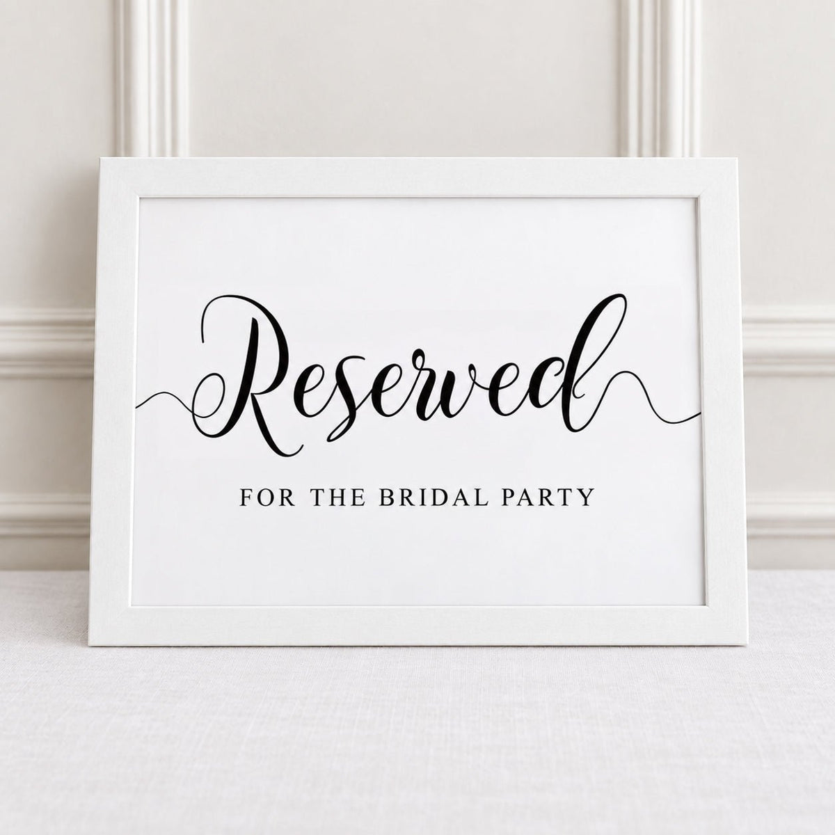 A white framed sign with black script reading 'Reserved' and smaller text 'FOR THE BRIDAL PARTY,' leaning against a light wall on a pale surface.