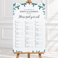 A vertical wedding reception seating chart printed on a white board with teal leafy watercolor accents at the top and bottom, displayed on a wooden easel in a bright indoor setting.