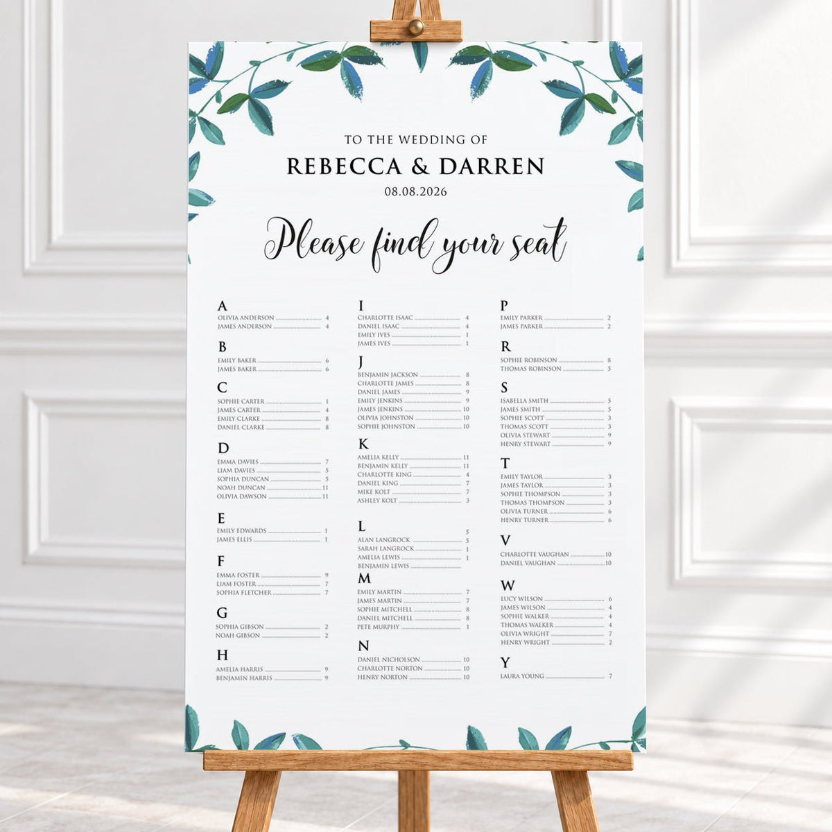 A vertical wedding reception seating chart printed on a white board with teal leafy watercolor accents at the top and bottom, displayed on a wooden easel in a bright indoor setting.