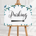 A rectangular white sign with the word "Parking" in black script and a black arrow pointing right, framed by blue-green leafy vines around the border. The sign is displayed on a wooden easel against a light interior wall.