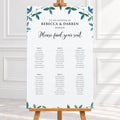 A wedding seating chart printed on a white card with teal leafy watercolor accents at the top and bottom, displayed on a wooden stand in a bright indoor setting.