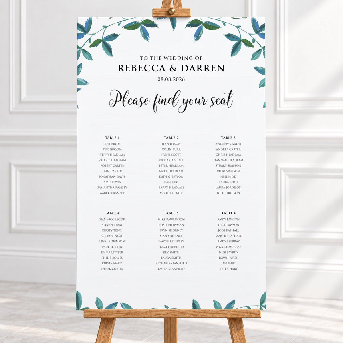 A wedding seating chart printed on a white card with teal leafy watercolor accents at the top and bottom, displayed on a wooden stand in a bright indoor setting.