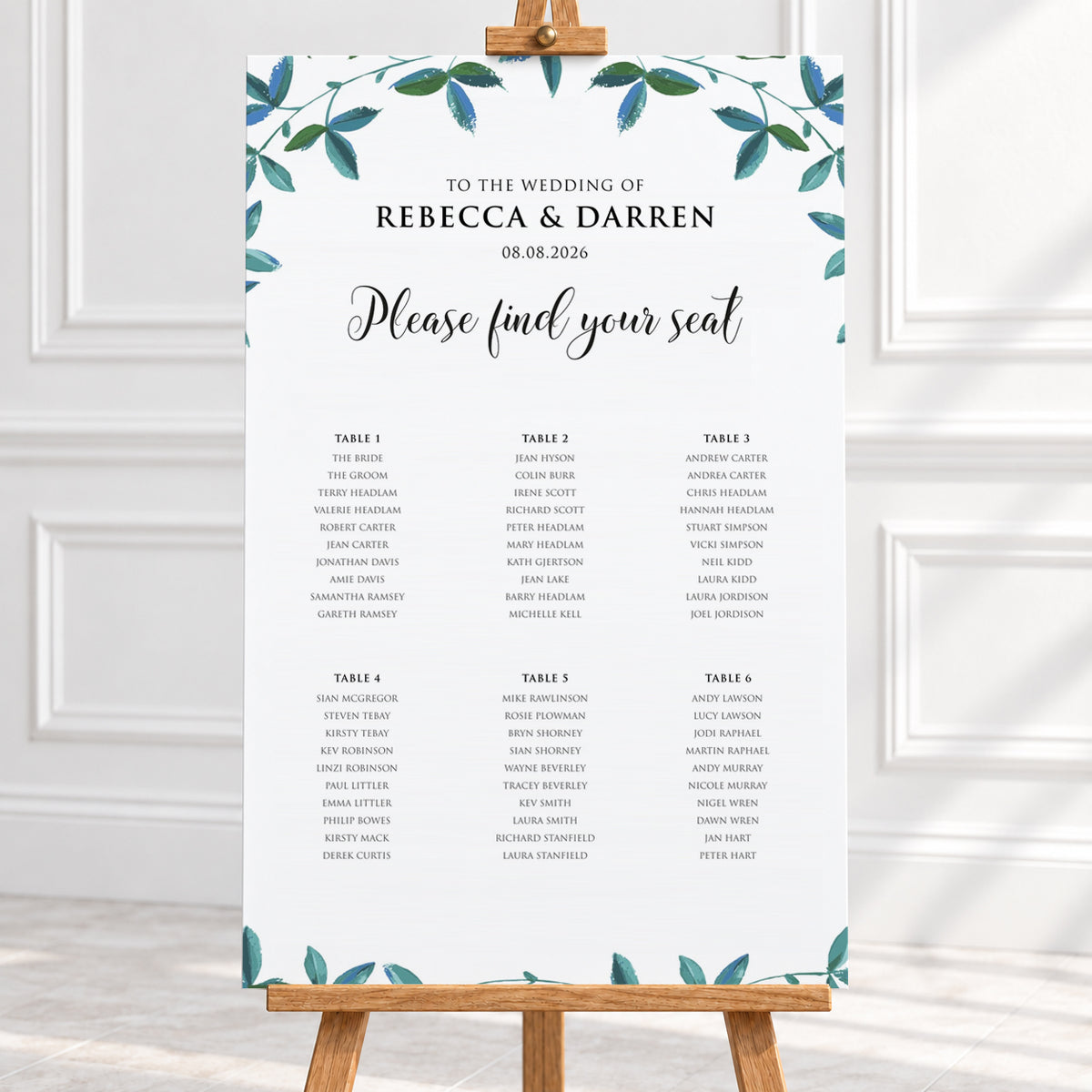 A wedding seating chart printed on a white card with teal leafy watercolor accents at the top and bottom, displayed on a wooden stand in a bright indoor setting.