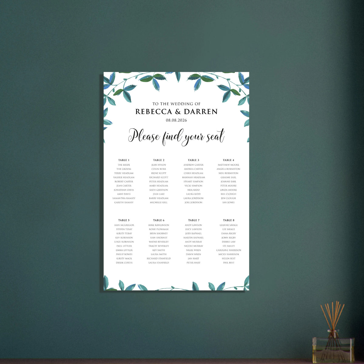 A wedding seating chart print with a white background, black text, and a teal and green leafy vine border, displayed against a dark green wall.