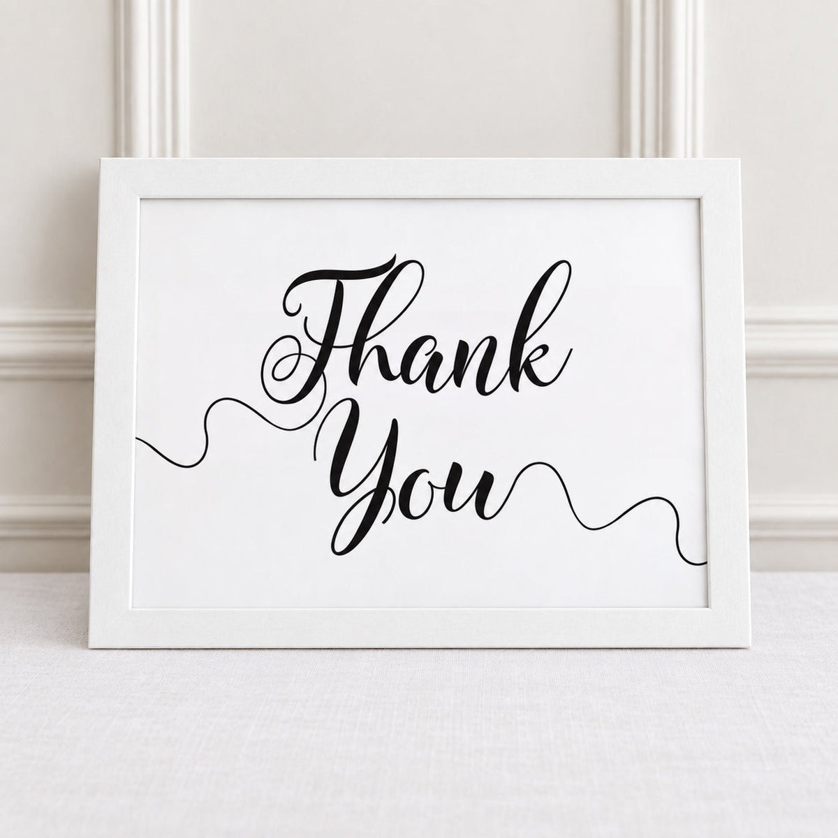 A white framed art print with the words 'Thank You' in elegant black cursive script, leaning against a white paneled wall on a light fabric surface.
