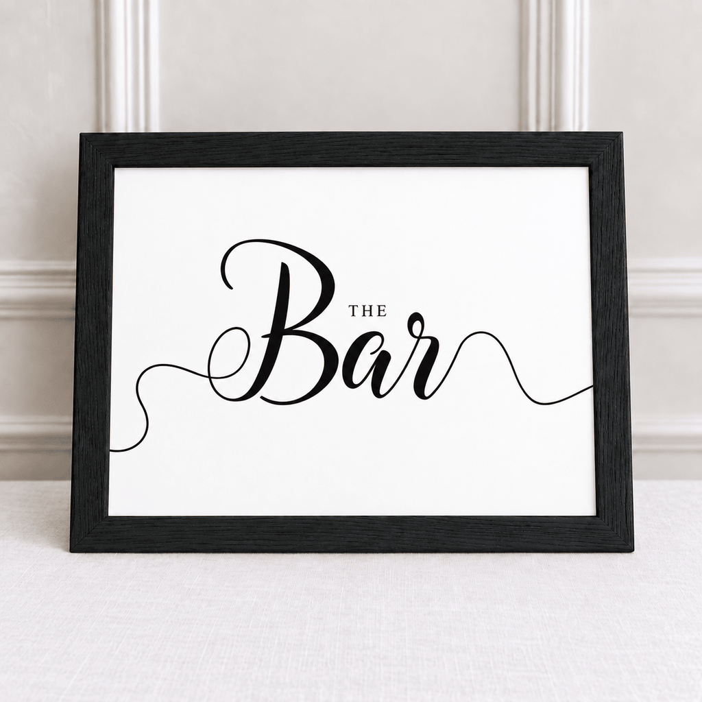 A framed typographic wall sign reading 'The Bar' in black script and small serif text, shown in a black frame against a light neutral wall and tabletop.