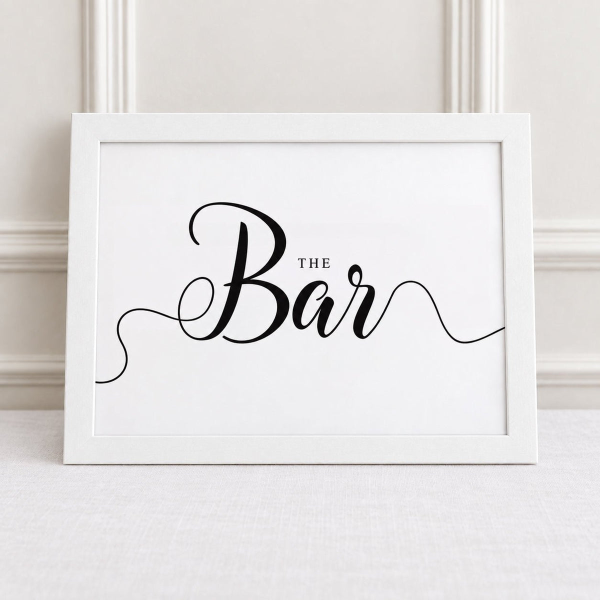 A white framed sign with the words 'THE Bar' in black script, leaning against a light wall on a pale surface.